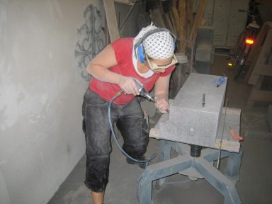 Kathrin Post-Isenberg arbeitet mit Staubmaske, Brille und Kopfbedeckung am staubigen Stein.