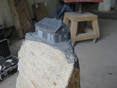Detailfoto eines Grabsteins, auf dem ein kleines Haus erarbeitet wurde.